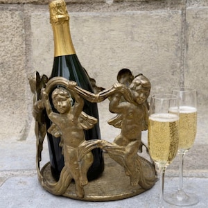 Puede incluir: Una botella de champán en un soporte decorativo de metal dorado con figuras de querubines. Dos copas de champán llenas de líquido burbujeante están cerca. La botella tiene una tapa de lámina dorada.