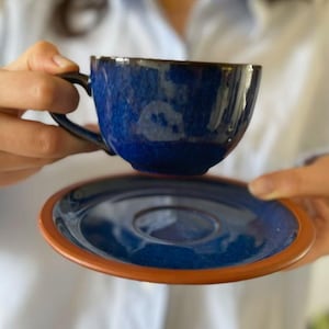 May include: A blue ceramic teacup and saucer set. The cup and saucer have a dark blue glaze with a slight iridescent sheen.