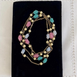 May include: A multi-strand necklace with faux pearls and colourful, irregular beads in pink, blue, and turquoise. The necklace is displayed on a black velvet surface, creating a striking contrast. The beads are in shades of pink, blue, and turquoise.