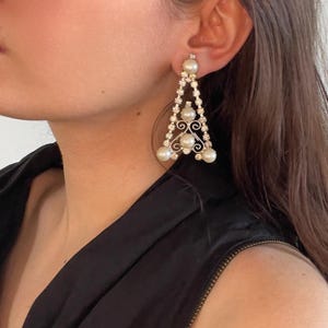 May include: A triangular drop earring featuring a silver-toned metal frame adorned with clear rhinestones and four round white pearls. The design includes a central scrollwork detail and a post-back closure, creating a geometric silhouette.