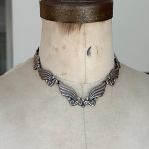 May include: A silver-toned metal choker necklace featuring a series of connected, wing-shaped links with curved, scroll-like details and small spherical accents. The necklace rests against a beige fabric mannequin neck form.