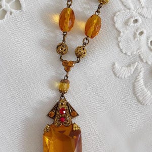 May include: Vintage amber-colored necklace with a rectangular pendant. The pendant is adorned with red and amber-colored accents. The necklace features a chain of amber-colored beads and ornate gold-toned metalwork.