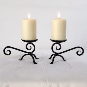 Puede incluir: Dos velas de pilar marfil encendidas sobre candelabros de hierro forjado negro. Los candelabros tienen un diseño en espiral y tres patas. Las velas están sobre un mantel blanco.