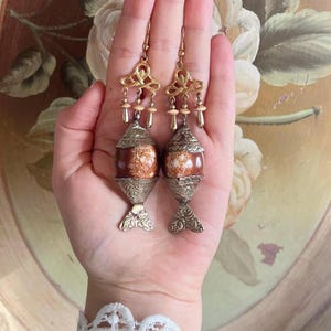 Puede incluir: Un par de pendientes ornamentados de estilo vintage. Los pendientes presentan una parte superior dorada con un diseño decorativo, pequeñas cuentas y un centro en forma de barril moteado marrón y blanco. La parte inferior de los pendientes tiene una base decorativa plateada.