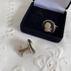 May include: A pair of gold-toned cufflinks with a black background and a white cameo profile. One cufflink is inside a black velvet box, and the other is lying on a white, embroidered surface. The cufflinks are a classic accessory.