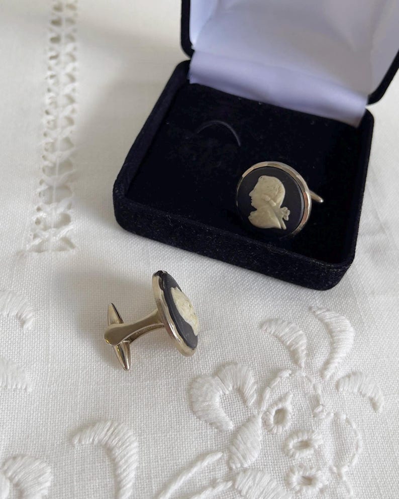 May include: A pair of gold-toned cufflinks with a black background and a white cameo profile. One cufflink is inside a black velvet box, and the other is lying on a white, embroidered surface. The cufflinks are a classic accessory.