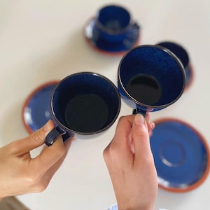Puede incluir: Dos tazas de café de cerámica azul con platillos a juego. Las tazas tienen un esmalte moteado y están sostenidas por dos manos.