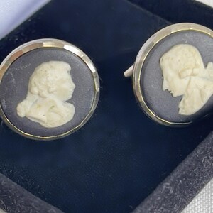 May include: A pair of cufflinks featuring a cameo design. Each cufflink has a silver-colored border surrounding a dark gray background with a cream-colored profile of a person. The cufflinks are presented in a black box.