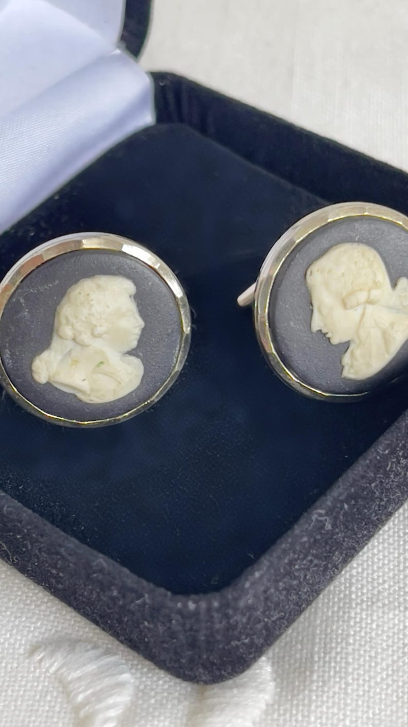 May include: A pair of cufflinks featuring a cameo design. Each cufflink has a silver-colored border surrounding a dark gray background with a cream-colored profile of a person. The cufflinks are presented in a black box.