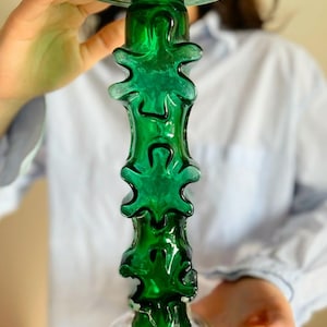 May include: A tall, green glass candle holder with a unique, textured design. The holder is made of thick glass and has a wide base.
