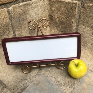 May include: A rectangular white sign with a dark maroon frame, supported by a gold-colored metal wire stand with decorative scrollwork at the top and base, positioned next to a single round yellow-green apple.