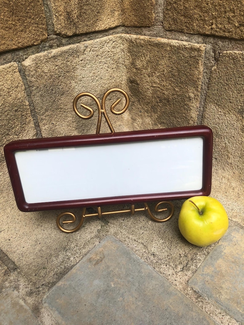 May include: A rectangular white sign with a dark maroon frame, supported by a gold-colored metal wire stand with decorative scrollwork at the top and base, positioned next to a single round yellow-green apple.