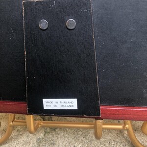 May include: The back of a rectangular picture frame with a black backing, a hinged support stand, and a white label that reads MADE IN THAILAND FAIT EN THAILANDE. The frame is resting on a gold-colored metal stand with curved hooks.