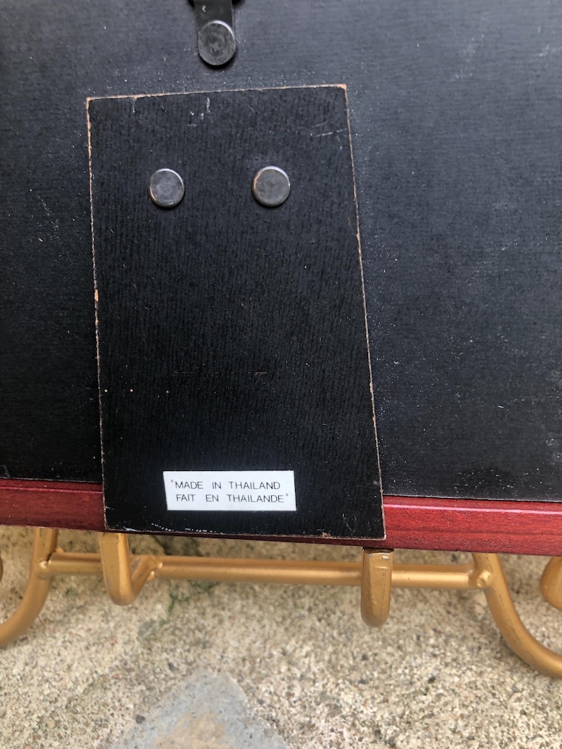 May include: The back of a rectangular picture frame with a black backing, a hinged support stand, and a white label that reads MADE IN THAILAND FAIT EN THAILANDE. The frame is resting on a gold-colored metal stand with curved hooks.