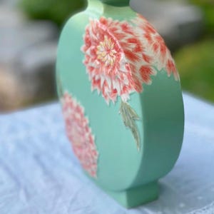 May include: A mint green ceramic vase with a flat, oval shape. The vase features two large, detailed floral designs in shades of red, pink, and yellow. The vase has a square top and base, and a height of approximately 10 inches.
