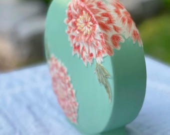 May include: A mint green ceramic vase with a flat, oval shape. The vase features two large, detailed floral designs in shades of red, pink, and yellow. The vase has a square top and base, and a height of approximately 10 inches.