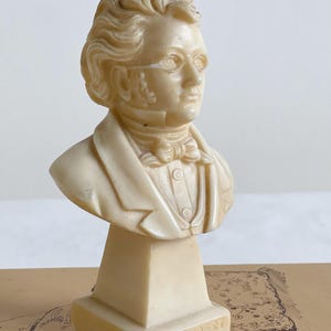 May include: A cream-colored bust of a man with glasses, a bow tie, and a jacket. The bust is mounted on a rectangular base with the name "SCHUBERT" inscribed. The sculpture is likely made of a composite material.