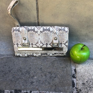 Op de afbeelding: Een rechthoekige polstas met een beige en bruin slangenleerpatroon. De tas heeft een polsband, een goudkleurige metalen balk aan de voorzijde en twee goudkleurige hardware-accenten. Een felgroene appel bevindt zich rechts van de tas.