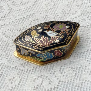 May include: A small, black decorative box with a hinged lid. The box is decorated with a gold floral and bird design. The box has a gold-coloured base and trim. The lid features a white bird perched on a branch with colourful flowers.