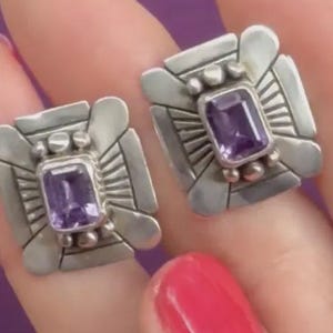 May include: A pair of square silver earrings featuring a central rectangular purple amethyst gemstone. The silver setting has a geometric design with radiating lines and small silver spheres surrounding the stone in a symmetrical pattern.