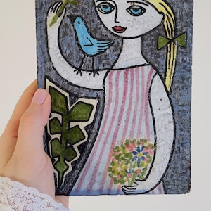 May include: A ceramic art piece depicting a girl with blonde hair, a pink and white striped dress, and a blue bird. The girl holds a flower and a bouquet of flowers. The background is a textured blue.