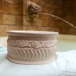 Puede incluir: Un cuenco redondo de cerámica de color claro con un patrón decorativo en relieve en el exterior. La sección superior muestra un diseño de volutas florales, mientras que la sección inferior está adornada con tallas estriadas verticales.