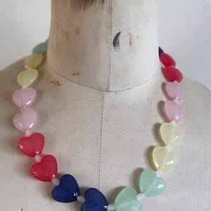 May include: A necklace featuring a series of heart-shaped beads arranged in a rainbow color gradient. The beads transition through shades of green, yellow, pink, red, and dark blue, strung together on a clear cord, creating a continuous circular pattern.