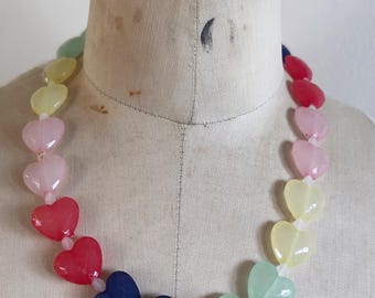 May include: A necklace featuring a series of heart-shaped beads arranged in a rainbow color gradient. The beads transition through shades of green, yellow, pink, red, and dark blue, strung together on a clear cord, creating a continuous circular pattern.