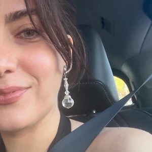 May include: A dangling earring featuring a clear, faceted crystal bead suspended from a smaller, elongated, diamond-shaped crystal component. The earring is attached to a silver-toned metal finding that secures it to the earlobe.