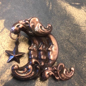May include: A copper-toned metal brooch featuring a crescent moon with three distinct faces in profile. The moon is adorned with ornate, swirling scrollwork details and has a small, five-pointed star charm hanging from the upper curve.