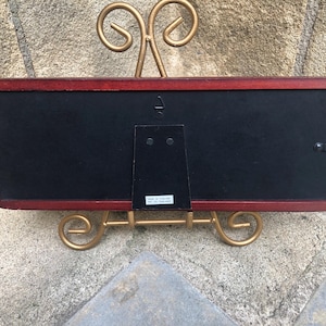 May include: A rectangular picture frame with a dark reddish-brown wooden border and a black backing. The frame features a central metal hanging hook and a hinged easel stand. A small white label on the back reads Made in Thailand.