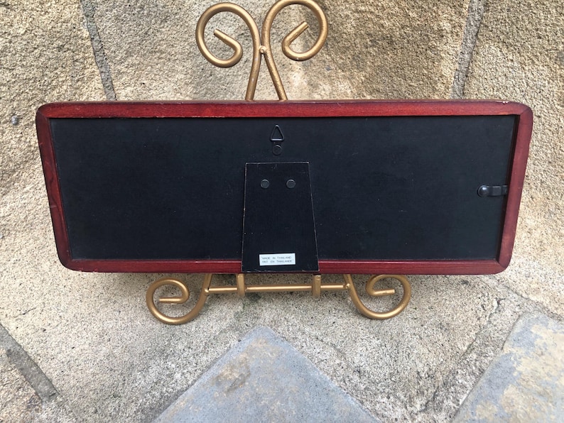 May include: A rectangular picture frame with a dark reddish-brown wooden border and a black backing. The frame features a central metal hanging hook and a hinged easel stand. A small white label on the back reads Made in Thailand.