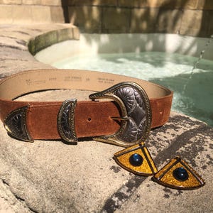 Könnte beinhalten: Ein brauner Wildledergürtel mit einer silber- und goldfarbenen Metallschnalle und passenden dekorativen Metallakzenten. Der Gürtel enthält eine Reihe von dreieckigen und halbmondförmigen Metallteilen mit eingravierten Mustern. Zwei separate dreieckige goldfarbene Ornamente mit dunkelblauen kreisförmigen Zentren liegen vor dem Gürtel.