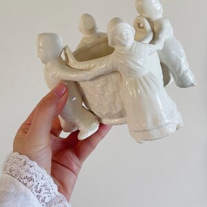 Puede incluir: Una maceta de cerámica blanca con un diseño de cesta, sostenida por cuatro figuras con túnicas. Las figuras parecen estar bailando o interactuando. La maceta se sostiene en una mano.