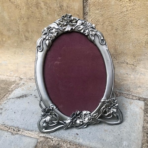 May include: An oval-shaped picture frame crafted from a silver-toned metal with a matte finish. The frame features raised, sculpted floral details at the top and bottom edges. The center of the frame contains a solid, dark burgundy velvet backing.