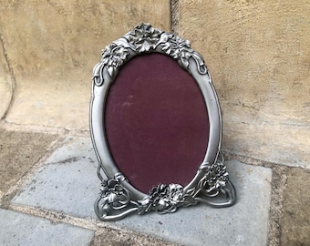 May include: An oval-shaped picture frame crafted from a silver-toned metal with a matte finish. The frame features raised, sculpted floral details at the top and bottom edges. The center of the frame contains a solid, dark burgundy velvet backing.