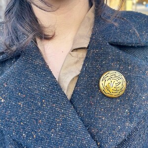 May include: A gold-colored, circular brooch with a unique, swirling design. The brooch is pinned to a dark blue tweed coat. The coat has a classic lapel and button closure. The overall aesthetic is vintage and elegant.