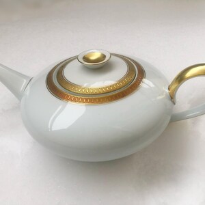 May include: White ceramic teapot with gold detailing. The teapot has a rounded shape, a curved handle, and a spout. The lid and handle are gold, and the body features a gold and brown decorative pattern. A stylish addition to any tea set.