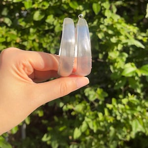 May include: A pair of clear, translucent hoop earrings with a smooth, rounded surface. The earrings feature a subtle, fine-textured mesh pattern embedded within the material, creating a semi-transparent appearance with a metallic sheen.
