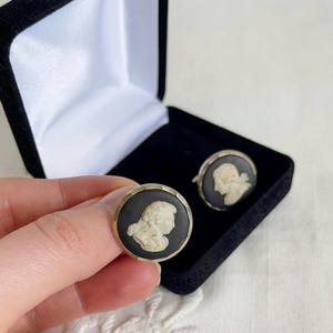 May include: A pair of vintage cufflinks featuring a black background with a cream-colored cameo profile. The cufflinks are round with a gold-colored border. One cufflink is held in a hand, and the other is in a black velvet box.