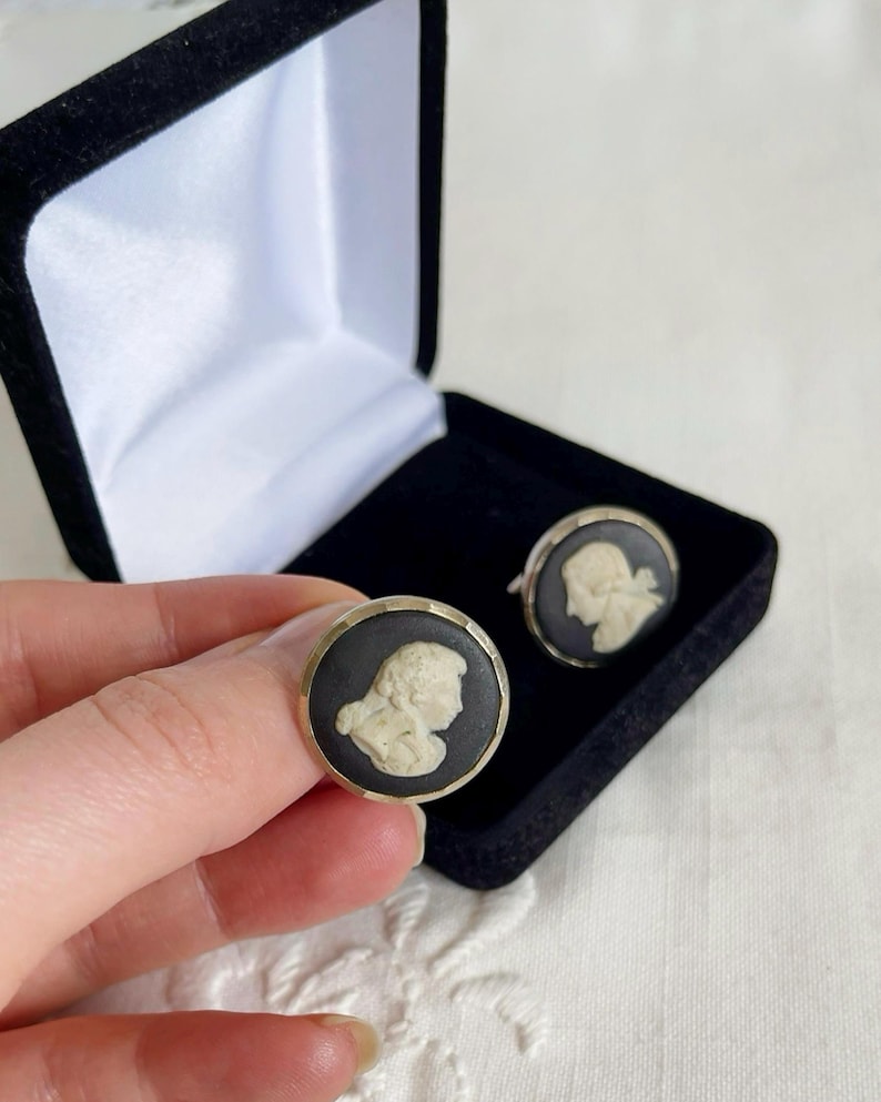 May include: A pair of vintage cufflinks featuring a black background with a cream-colored cameo profile. The cufflinks are round with a gold-colored border. One cufflink is held in a hand, and the other is in a black velvet box.