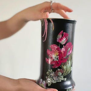 May include: A cylindrical black vase featuring a hand-painted floral design with vibrant pink and red flowers, green foliage, and delicate pink trailing accents near the top rim. The vase has a smooth matte finish and a slightly flared base.
