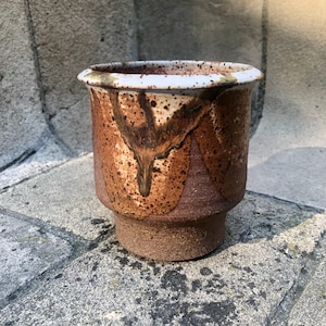 May include: A ceramic planter with a wide, flared rim and a tapered base. The exterior features a textured, unglazed brown clay bottom section, while the upper portion is finished with a speckled, drip-style glaze in shades of cream and dark brown.