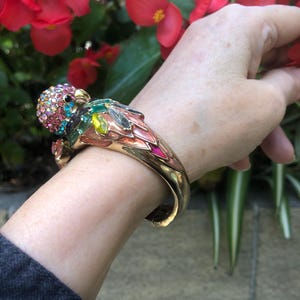 May include: A gold-tone hinged bangle bracelet shaped like a parrot. The bird head is covered in pink, blue, and clear rhinestones. The wings feature marquise-cut green, yellow, and clear crystals with pink and gold enamel detailing on the metal.