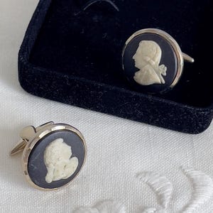 May include: A pair of vintage cufflinks with a black background and a white cameo profile design. The cufflinks are round with a gold-colored metal border and are displayed in a black velvet box.