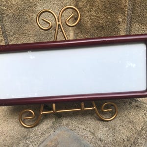 May include: A rectangular, dark maroon-colored frame with a smooth, rounded edge finish. The frame features a blank, white, flat rectangular center panel. It is supported by a gold-toned metal display stand with decorative, curved scrollwork feet.
