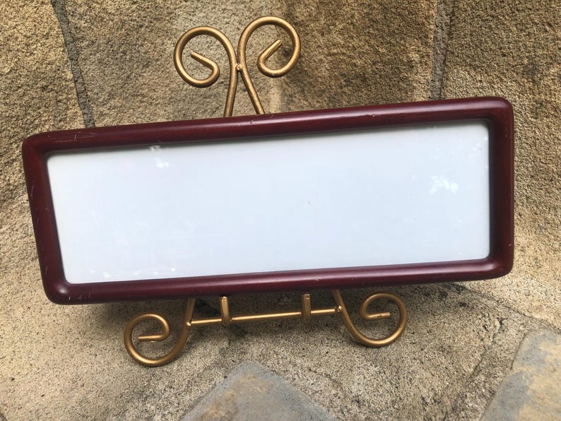 May include: A rectangular, dark maroon-colored frame with a smooth, rounded edge finish. The frame features a blank, white, flat rectangular center panel. It is supported by a gold-toned metal display stand with decorative, curved scrollwork feet.