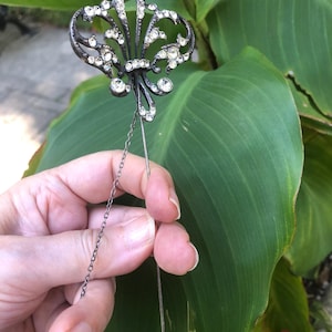 May include: A dark metal lapel pin featuring a fleur-de-lis design encrusted with clear, sparkling rhinestones. The pin includes a long, slender metal stem and a delicate, thin chain attached to the base of the decorative top element.