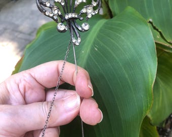May include: A dark metal lapel pin featuring a fleur-de-lis design encrusted with clear, sparkling rhinestones. The pin includes a long, slender metal stem and a delicate, thin chain attached to the base of the decorative top element.