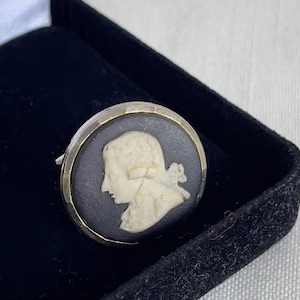 May include: A vintage cameo ring featuring a cream-colored profile set against a black background. The ring has a silver-colored metal band and is displayed in a black velvet box. The cameo depicts a classical figure.
